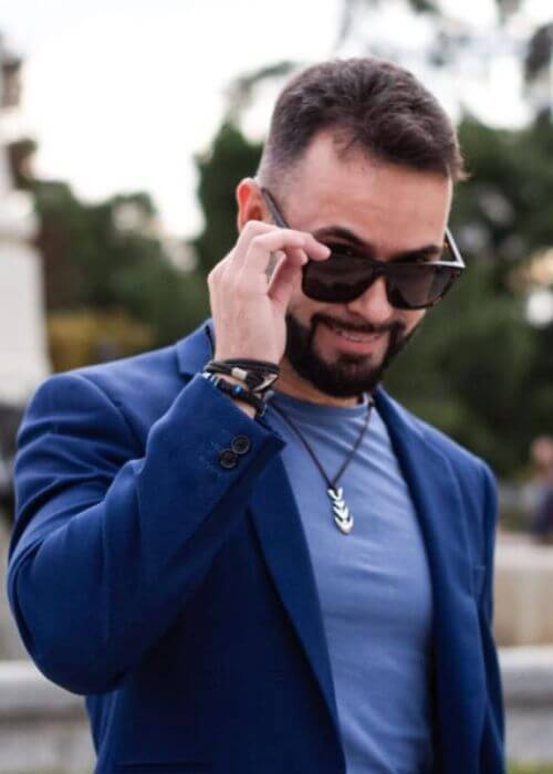 Antonio Gallo, con un blazer azul y gafas de sol, mira hacia abajo, ajustándose las gafas contra un fondo exterior borroso.