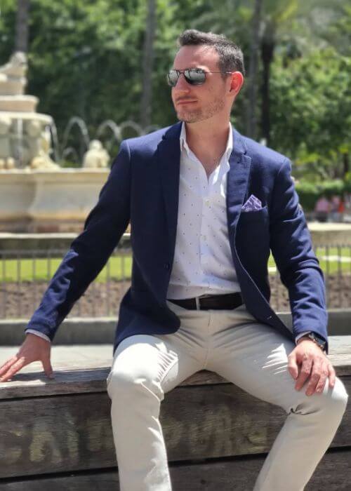 Antonio Parra, con gafas de sol y un blazer azul, se sienta en un banco con una fuente y árboles al fondo.