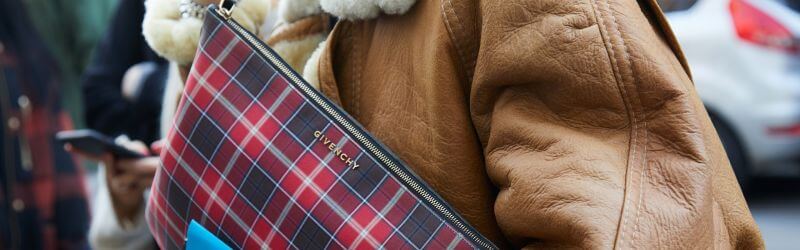 Persona con chaqueta de piel de oveja marrón que lleva un bolso de mano Givenchy de cuadros rojos; se ven coches en el fondo borroso.