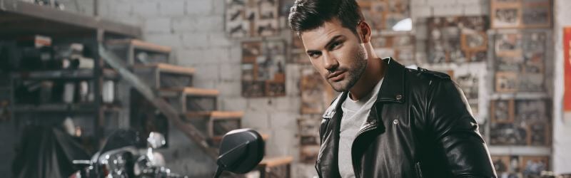 Hombre con chaqueta de cuero negra junto a una motocicleta en un garaje con paredes de ladrillo y escaleras de madera al fondo.
