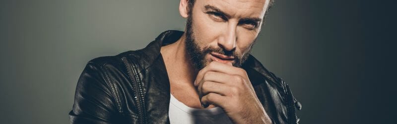 Hombre con barba vestido con chaqueta de cuero negra y camisa blanca, mirando a la cámara con la mano en la barbilla.