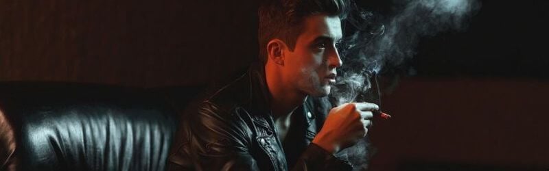 Un hombre con chaqueta de cuero se sienta en un sofá oscuro, sosteniendo un cigarrillo cuyo humo se eleva en la penumbra.