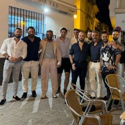 Un grupo de diez hombres de pie juntos al aire libre, sonriendo por la noche, con un bar y edificios visibles al fondo.