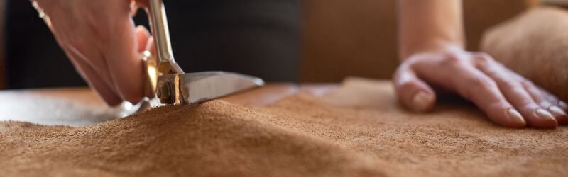 Primer plano de una persona cortando tela de ante marrón con tijeras metálicas sobre una mesa de trabajo.