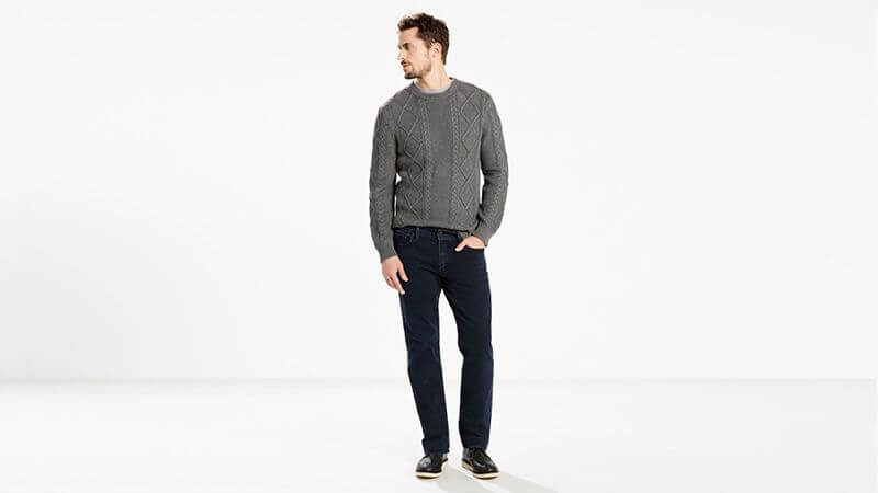 Hombre con jersey gris, vaqueros oscuros y zapatos negros de pie en un estudio blanco, mirando a su izquierda.