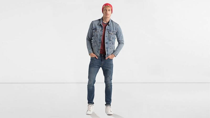 Hombre vestido con chaqueta vaquera, vaqueros, gorro de punto rojo y zapatillas de deporte blancas sobre un fondo blanco liso.