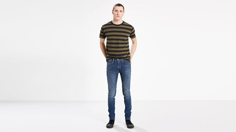 Una persona de pie, mirando al frente, lleva una camiseta de rayas, vaqueros azules y zapatos negros sobre un fondo liso.