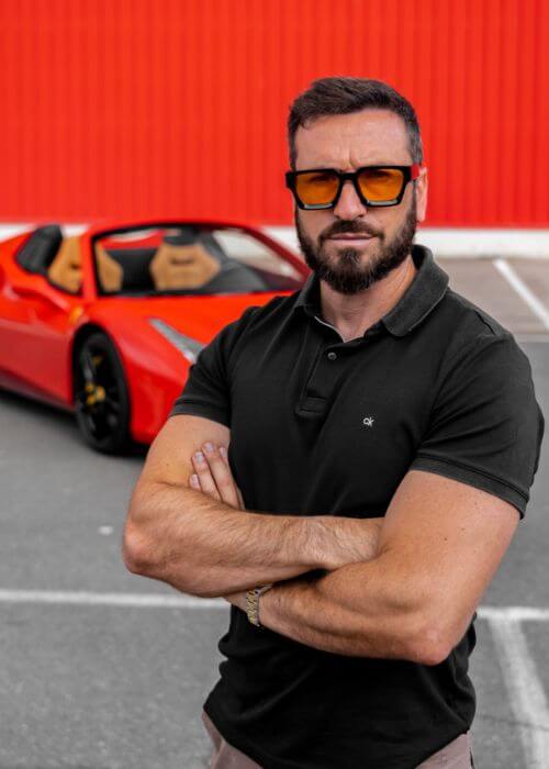 Manu Solís con gafas de sol y los brazos cruzados se encuentra frente a un Ferrari deportivo rojo en un estacionamiento con un fondo de pared naranja.