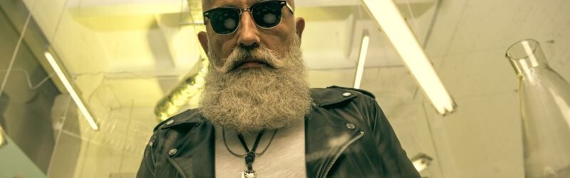 Un hombre con una espesa barba gris y gafas de sol que lleva una chaqueta de cuero permanece de pie en un interior bajo luces fluorescentes.